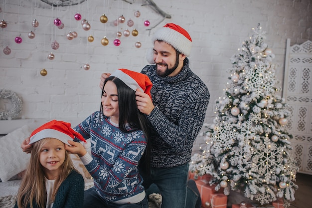 Фото Веселого рождества и счастливых праздников веселая мама, папа и ее милая дочь одевают друг другу рождественские шляпы. родитель и маленький ребенок с удовольствием возле елки в помещении. утро рождество.