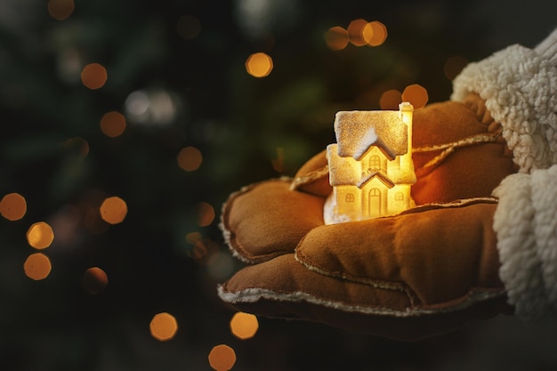 写真 メリー クリスマスとハッピー ホリデー ライトアップされたクリスマス ツリー ライトの背景に小さな輝く家を保持している手袋で手魔法の冬時間大気イメージ居心地の良い家