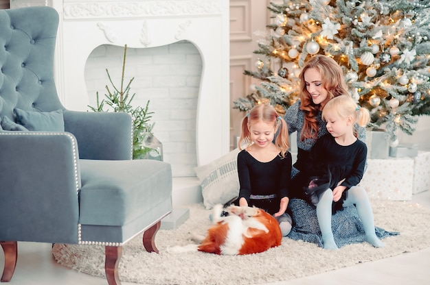 사진 즐거운 성탄절과 즐거운 휴일 보내세요. 엄마와 아이들은 개를 쓰다듬고, 딸들은 실내 나무 근처에서 즐거운 시간을 보냅니다. 방에서 사랑하는 가족.