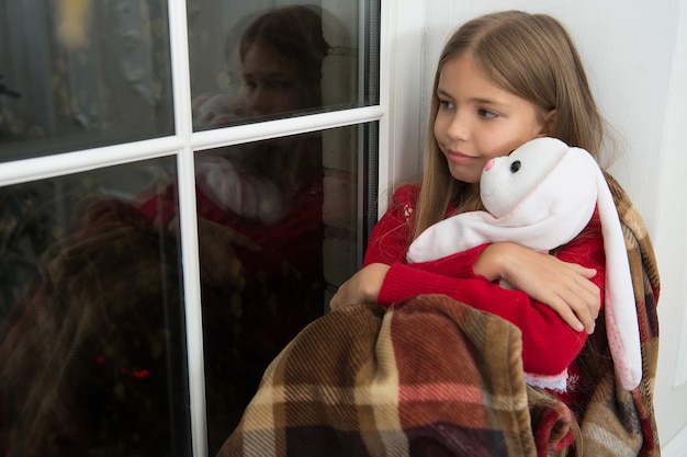 Фото Веселого рождества и счастливого нового года. маленький ребенок с игрушкой кролик у окна. маленькая девочка держит игрушечного друга. маленький ребенок расслабиться в канун рождества. маленькая девочка наслаждается рождественским духом дома. единение.