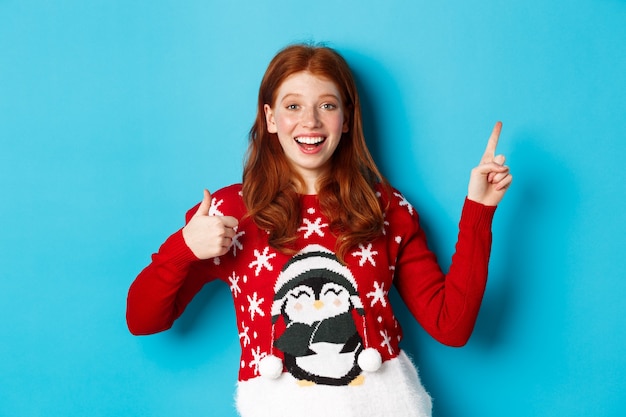 Merry Christmas. Cheerful redhead girl in xmas sweater, pointing finger at upper right corner, showing new year promo and thumbs-up in approval, praise product