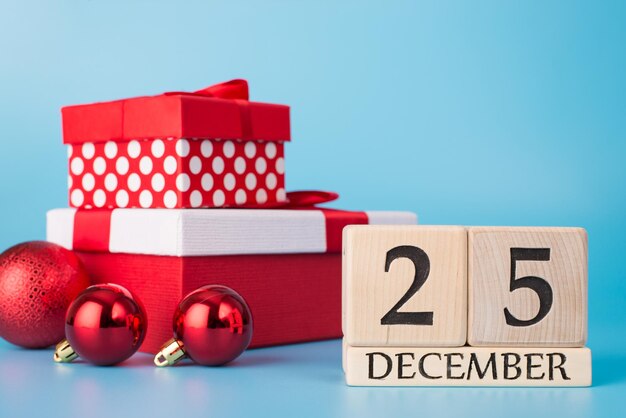 merry christmas concept. close up photo of wooden block calendar showing 25th of december and pile of red presents balls isolated over blue background