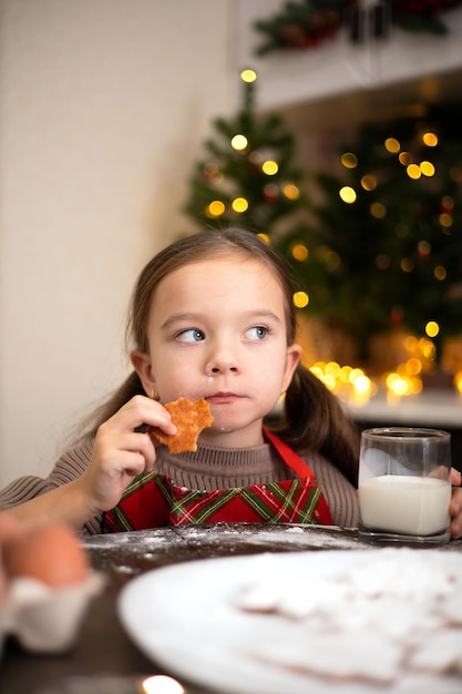 Счастливого Рождества Забавная милая девочка на уютной украшенной кухне печет рождественское печенье и ест его Стиль жизни Теплые тона