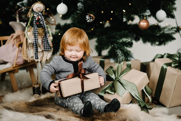 Merry Christmas and Happy Holidays Cheerful cute child girl opening gifts Kid having fun near tree in the morning Loving family with presents in room