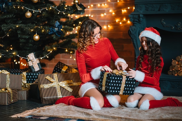 メリークリスマスとハッピーホリデー！陽気なお母さんとクリスマスの衣装を着たかわいい娘の女の子がプレゼントを交換します。