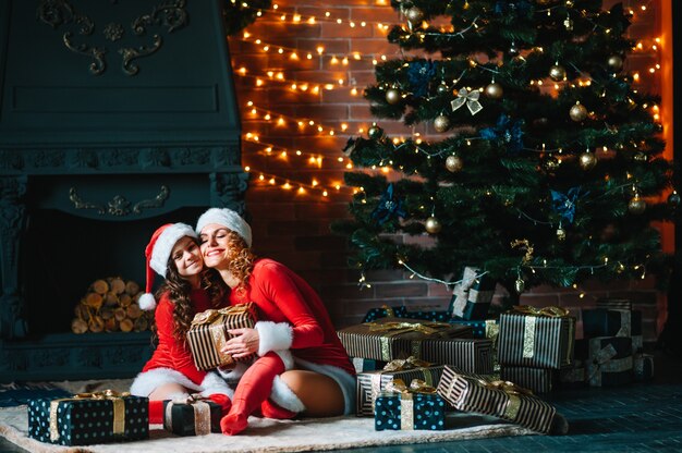 メリークリスマスとハッピーホリデー！陽気なお母さんとクリスマスの衣装を着たかわいい娘の女の子がプレゼントを交換します。