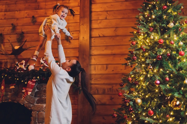 メリークリスマスとハッピーホリデー陽気なママとかわいい娘の女の子が贈り物を交換します。親と小さな子供が室内のクリスマスツリーの近く楽しんで。部屋にプレゼントがある愛情のある家族。