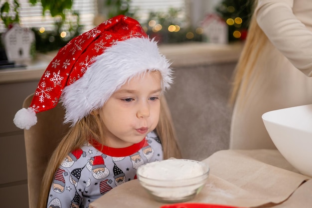 Merry Christmas and happy holidays. A cute girl in Santa's hat and pajamas in the kitchen decorated for Christmas plays with flour High quality photo