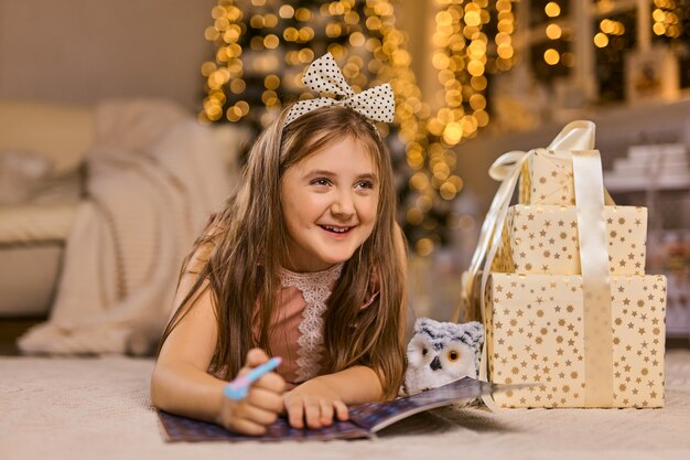 メリークリスマスとハッピーホリデー。かわいい小さな子供の女の子は、屋内のクリスマスツリーの近くのサンタクロースに手紙を書きます。