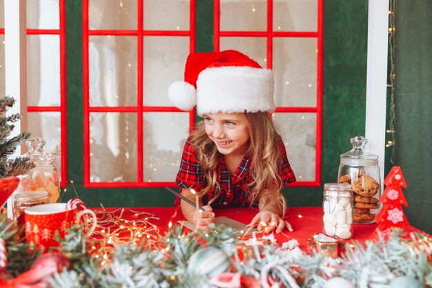 Merry christmas and happy holidays a cute little girl in\
pajamas writes a letter to santa claus near the christmas tree\
indoors children