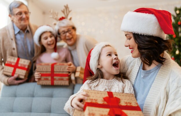 メリークリスマスとハッピーホリデー！おばあちゃん、おじいちゃん、お母さんと子供たちが贈り物を交換します。屋内の木の近くで楽しんでいる親と娘。部屋にプレゼントを持っている愛する家族。