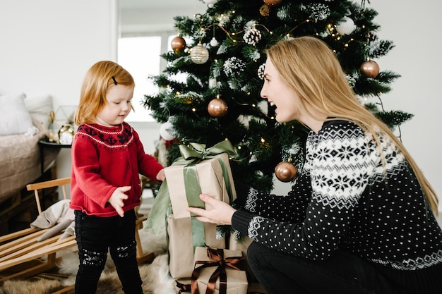 メリークリスマスとハッピーホリデー ママはクリスマスツリーの近くで娘にプレゼントを贈る 陽気な母親が家で一緒にかわいい赤ちゃんの女の子を抱きしめる 新年あけましておめでとうございます2021