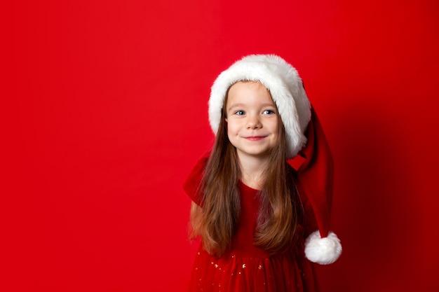 メリークリスマスと幸せな休日赤い背景の上のサンタの帽子の感情的な女の子の肖像画