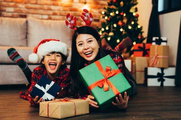 Merry christmas and happy holidays! young elegant mom and cute
child received many christmas gift on boxing day showing to camera
cheerful smiling. sweet family lying on wooden floor in warm
house.