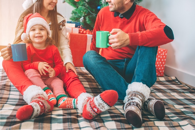 Merry Christmas and Happy New Year. Cut view of man and woman sit on blanket with their child. They hold cups and look at each other. They smile. Kid look and camera and laugh.