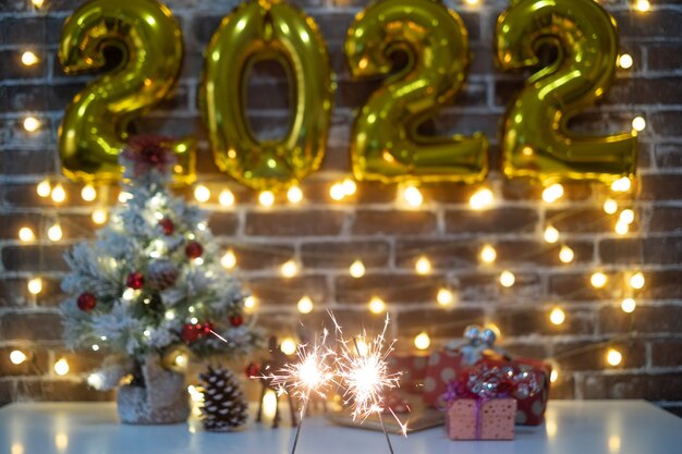 Photo merry christmas and happy new year, defocused small christmas tree on table with christmas presents and 2022 in golden inflatable numbers. sparkling lights. brick wall background