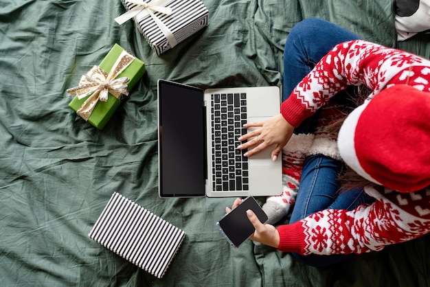 즐거운 성탄절 보내시고 새해 복 많이 받으세요. 온라인 쇼핑. 노트북을 사용하여 침대에 앉아 크리스마스 옷을 입고 상위 뷰 여자. 모형 화면, 복사 공간