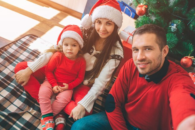 メリークリスマス、そしてハッピーニューイヤー。素敵な家族の写真。若い男はカメラを持って自分撮りをします。それらのすべてがポーズします。子供は真面目そうだ。