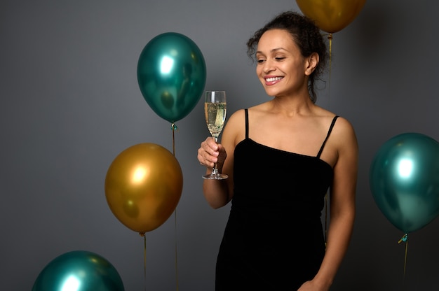 Merry Christmas and Happy New Year. Promotional shot of a beautiful Hispanic woman in evening dress with glass of champagne posing against gray background with shiny gold and green metallic balloons