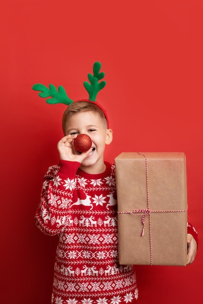 Merry Christmas and Happy New Year. Studio shot of Caucasian happy boy wearing reindeer antlers, holding gift box, isolated on red background.