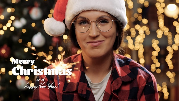 Merry Christmas and happy New Year text. Beautiful woman smiles and looks into camera, sparkler in her hand, light of garland and sparkler is reflected in glasses. Close-up