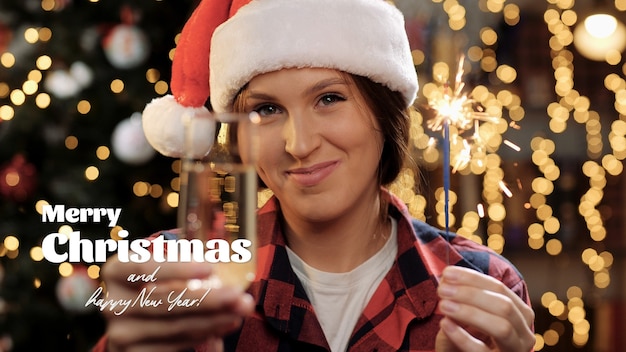 Merry Christmas and happy New Year text. Happy woman smiles and looks at camera, she holding sparkler and glass of champagne in hand, she makes toast gesture. Close-up