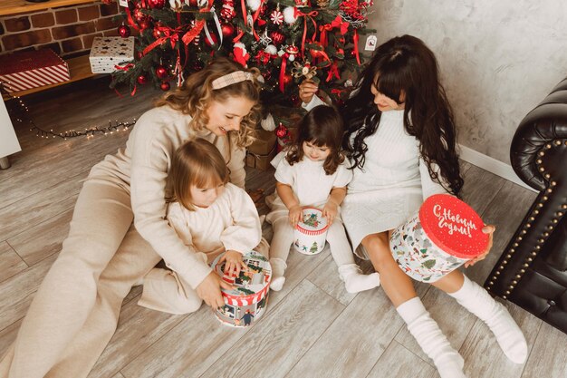 Merry Christmas and Happy New Year! Two women with their young daughters are sitting on the floor near the Christmas tree and opening presents.