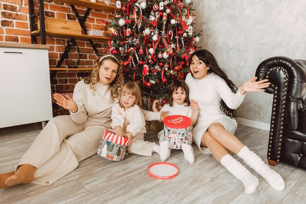 メリークリスマス、そしてハッピーニューイヤー！ 2人の若い娘を持つ女性がクリスマスツリーの近くの床に座ってプレゼントを開けています。