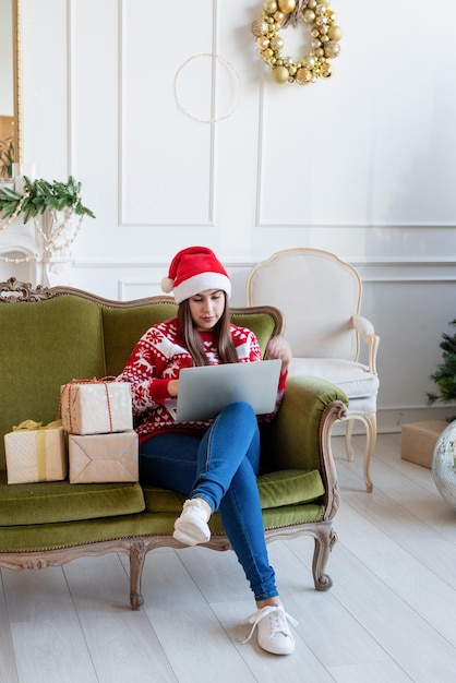Веселого Рождества и счастливого Нового года. Молодая брюнетка в шляпе санта-клауса сидит на зеленом диване и работает над ноутбуком в украшенной гостиной