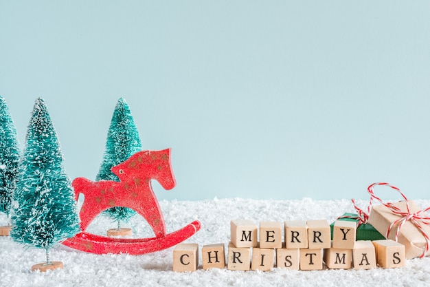 Photo merry christmas inscription with fir trees, horse toy, gift boxes on snow background