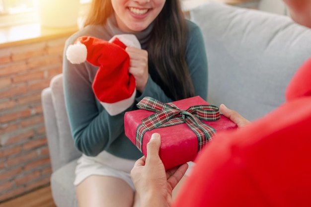 Buon natale. adorabile giovane coppia romantica sorprendente e regala una confezione regalo di natale alla sua ragazza seduta sul divano che festeggia il natale in casa, vacanze, felice anno nuovo e concetto di festival