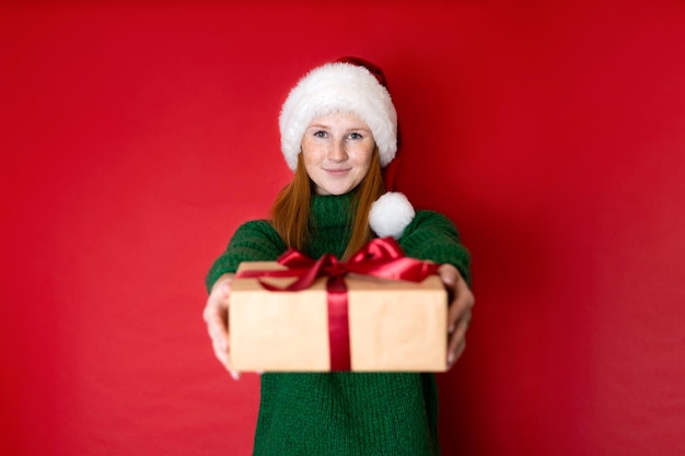 Merry christmas portrait of a beautiful young teenage girl in a\
cozy knitted green sweater and santa39s hat holding gift boxes the\
red background is the place for the text