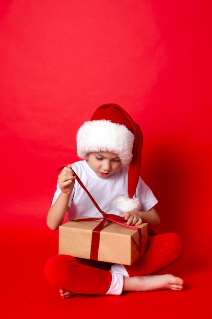 メリークリスマス。赤い背景の上のギフトボックスとサンタの帽子のかわいい男の子の肖像画。テキストの場所。高品質の写真