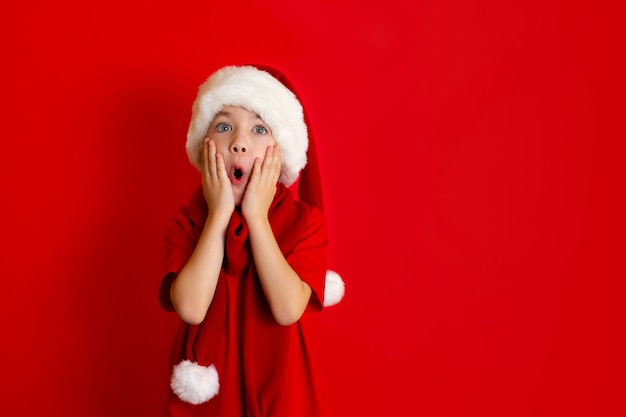 メリークリスマス。赤い背景の上の赤いTシャツのクリスマスキャップで面白い陽気な男の子の肖像画。テキストの場所。高品質の写真