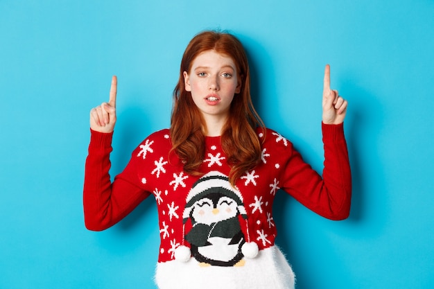 Merry christmas. skeptical and unamused redhead girl pointing fingers up, showing logo with reluctant face, standing over blue background