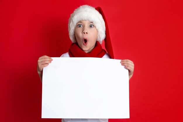 Счастливого Рождества. Сюрприз. Портрет забавного мальчика в новогодней шапке с белым листом в руках на красном фоне. место для текста. Эмоции. Фото высокого качества