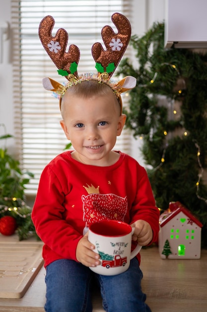 Merry Christmas Sweet little boy sits in the kitchen decorated for Christmas with a mug of eggnog drink. Traditions. High quality photo