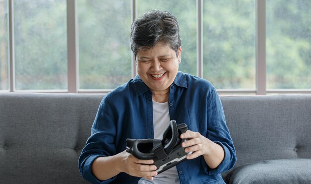 Merry mature Asian woman smiling and inspecting modern VR glasses while sitting on couch near window at home