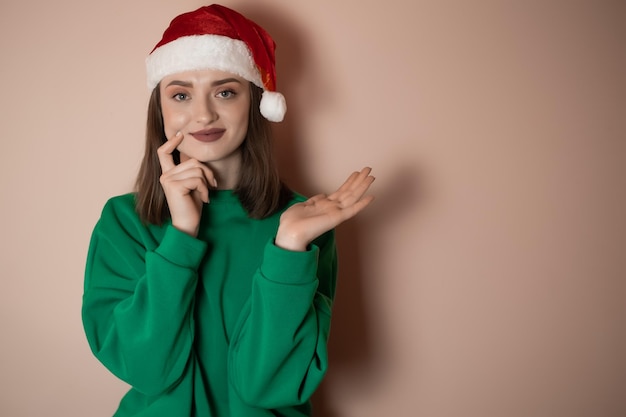 Merry surprised young woman wear xmas sweater santa hat posing\
pointing hands arms aside indicate on workspace area isolated on\
plain pastel light green background happy new year 2023 holiday\
concept