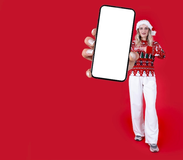 Merry young full body woman in knitted christmas sweater and santa hat hold big mobile phone mock up