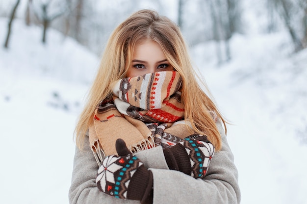冬の雪の公園でヴィンテージミトンのスタイリッシュな暖かい灰色のコートを着た驚くべき茶色の目を持つ陽気な若い幸せな女性。彼女の顔にウールのスカーフを持つ陽気なスタイリッシュな女の子。