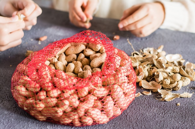 Mesh bag with inshell peanuts and two girls in the background are peeling nuts at the table. Lifestyle