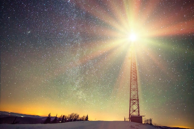 Mesmerizing landscape of the starry night sky with a radiant satellite tower spread over a snow-covered hill at the top of the mountain. Concept of weather station and beauty of the northern nature