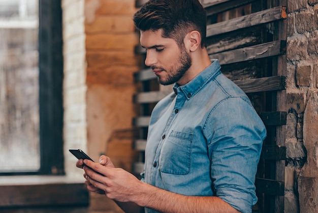 Message to her.Side view of handsome thoughtful young man holding smart phone and looking at it while leaning to brick wall in loft interior