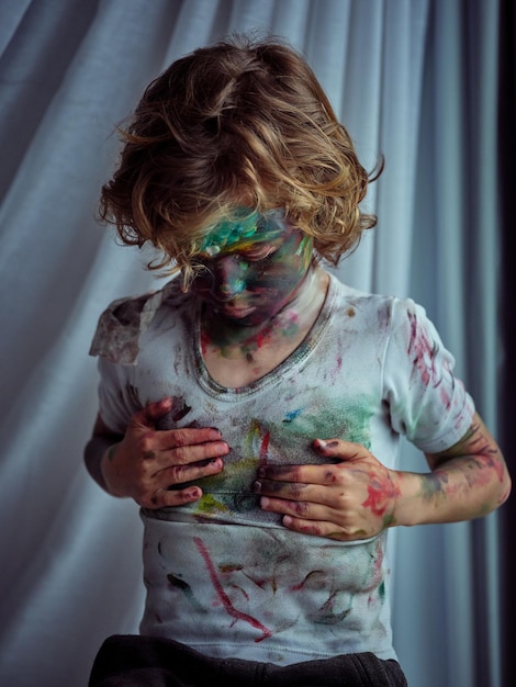 Foto ragazzo disordinato con i capelli ricci e il viso dipinto che si asciuga le mani sporche su una maglietta disordinata mentre si trova in una stanza luminosa