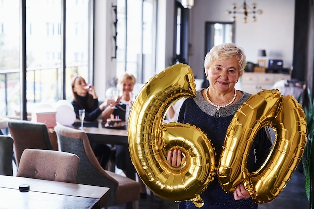 Met ballonnen van nummer 60 in handen. senior vrouw met familie en vrienden die een verjaardag binnenshuis viert.