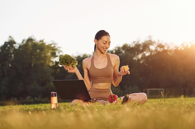 Met behulp van laptop Holding groenten Jonge vrouw in yoga kleding is buiten op het veld