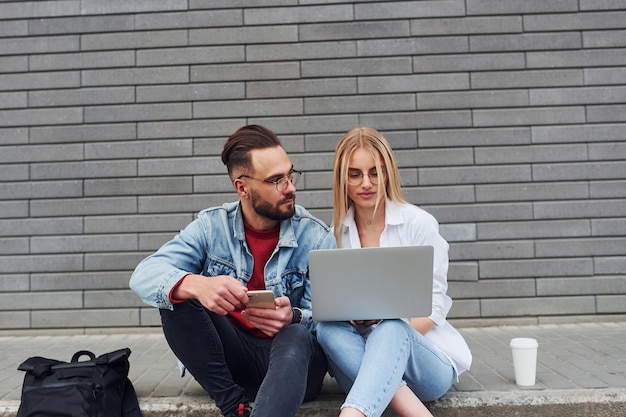 Met behulp van laptop Stijlvolle jongeman met vrouw in vrijetijdskleding die samen buiten zit Conceptie van vriendschap of relaties