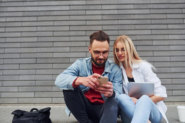 Met behulp van laptop stijlvolle jongeman met vrouw in vrijetijdskleding die samen buiten zit conceptie van vriendschap of relaties