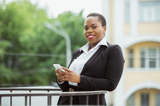 Met behulp van smartphone Afro-Amerikaanse zakenvrouw in kantoorkleding glimlachend ziet er zelfverzekerd en gelukkig uit